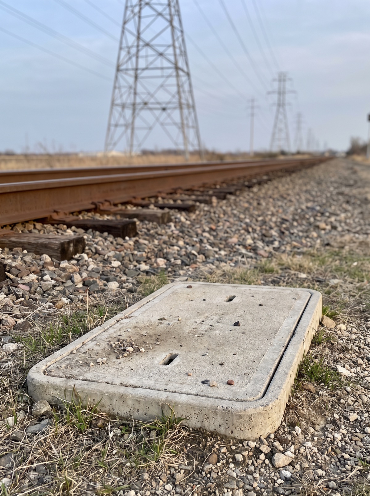 At-grade concrete chamber cover beside a railroad track with high-voltage power lines overhead, the type of site where the Merlin reads stray current